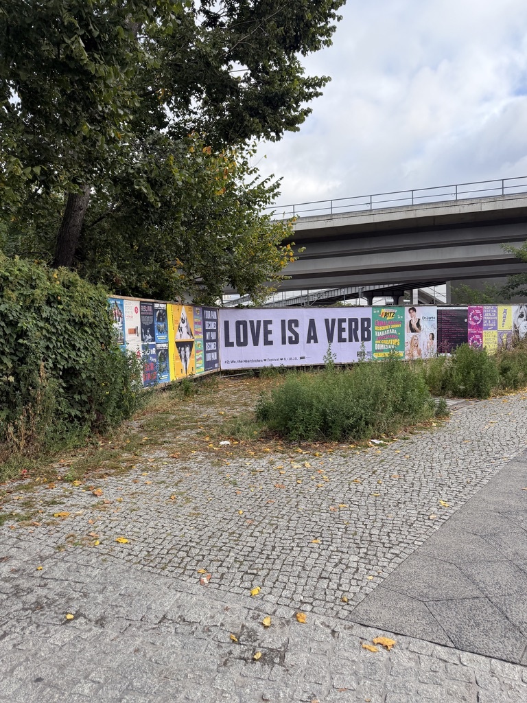 a 'love is a verb' billboard on a poster-covered fence next to a cobblestone path and a highway overpass, a few yellow leaves on the ground